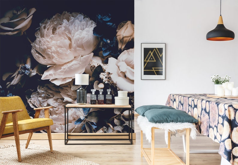 K&ouml;nnte beinhalten: Eine florale Tapeten-Wandmalerei mit gro&szlig;en wei&szlig;en Pfingstrosen und blauen Blumen auf einem dunklen Hintergrund. Die Wandmalerei ist in einem Wohnzimmer mit einem gelben Sessel, einem Holz-Konsolentisch und einer schwarzen Pendelleuchte dargestellt.