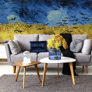 Puede incluir: Un sofá gris con una manta negra y un cojín de terciopelo azul se encuentra frente a un mural de pared que representa un cielo nocturno estrellado con pájaros volando sobre un campo de trigo. Una mesa de café blanca con un jarrón de flores amarillas se encuentra frente al sofá.