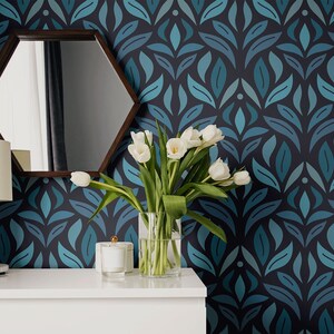Puede incluir: Un espejo hexagonal blanco con un marco de madera oscura cuelga de una pared con un papel tapiz de patrón floral azul teal y azul marino. Una lámpara blanca con una pantalla blanca se encuentra sobre una mesa blanca frente al espejo. Un jarrón de vidrio transparente con tulipes blancas se encuentra sobre la mesa.