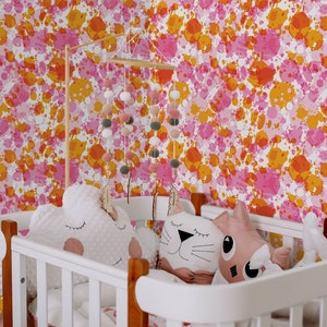 May include: A white crib with a pink and orange splatter paint wallpaper. The crib has a white wooden frame and a white mattress. There are two stuffed animals in the crib, a white cloud-shaped one and a pink cat-shaped one.