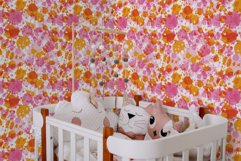 May include: A white crib with a pink and orange splatter paint wallpaper. The crib has a white wooden frame and a white mattress. There are two stuffed animals in the crib, a white cloud-shaped one and a pink cat-shaped one.