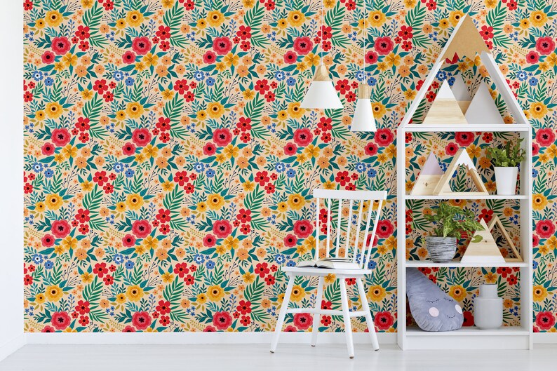 May include: A white bookshelf with three shelves, decorated with wooden geometric shapes and potted plants, against a wall covered in a floral wallpaper with red, yellow, and blue flowers on a cream background.