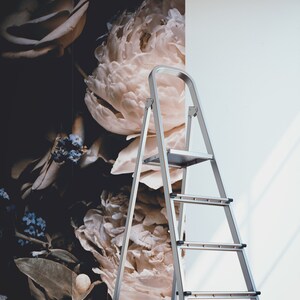 K&ouml;nnte beinhalten: Eine silberne Trittleiter mit drei Stufen steht vor einer Wand mit einer floralen Tapete mit gro&szlig;en wei&szlig;en Pfingstrosen und Rosen. Zwei Rollen Tapeten liegen auf dem Boden.