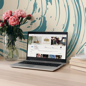 May include: A laptop computer open to an Etsy shop website with a product listing for a gold metal sign that says "It's a wonderful life". The laptop is on a light wood desk with a vase of pink peonies, a white coffee mug, and a stack of books.