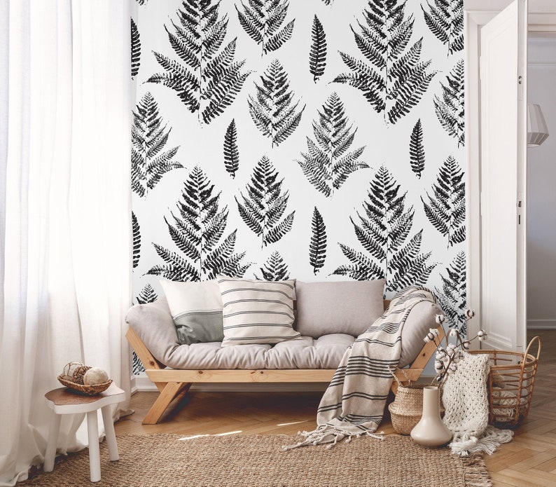 K&ouml;nnte beinhalten: Ein Wohnzimmer-Interieur mit einer wei&szlig;en Wand, die mit einer schwarz-wei&szlig;en Farn-Mustertapete versehen ist. Vor der Wand steht ein hellgraues Sofa mit Kissen und einer Decke. Im Vordergrund steht ein kleiner Holztisch mit einer Schale voller Dekoartikel.