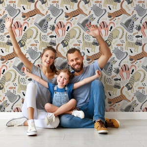 May include: A family of three poses in front of a wall with a whimsical wallpaper featuring a repeating pattern of colorful cats and fish. The wallpaper is a light beige with brown, pink, and gray cats.