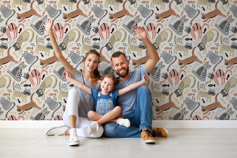 May include: A family of three poses in front of a wall with a whimsical wallpaper featuring a repeating pattern of colorful cats and fish. The wallpaper is a light beige with brown, pink, and gray cats.