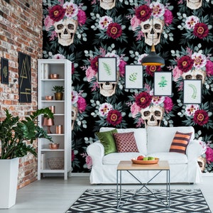 May include: A white couch with green and red pillows sits in front of a black and white floral wallpaper with skulls. The wallpaper features pink and red flowers, green leaves, and white skulls with floral crowns.