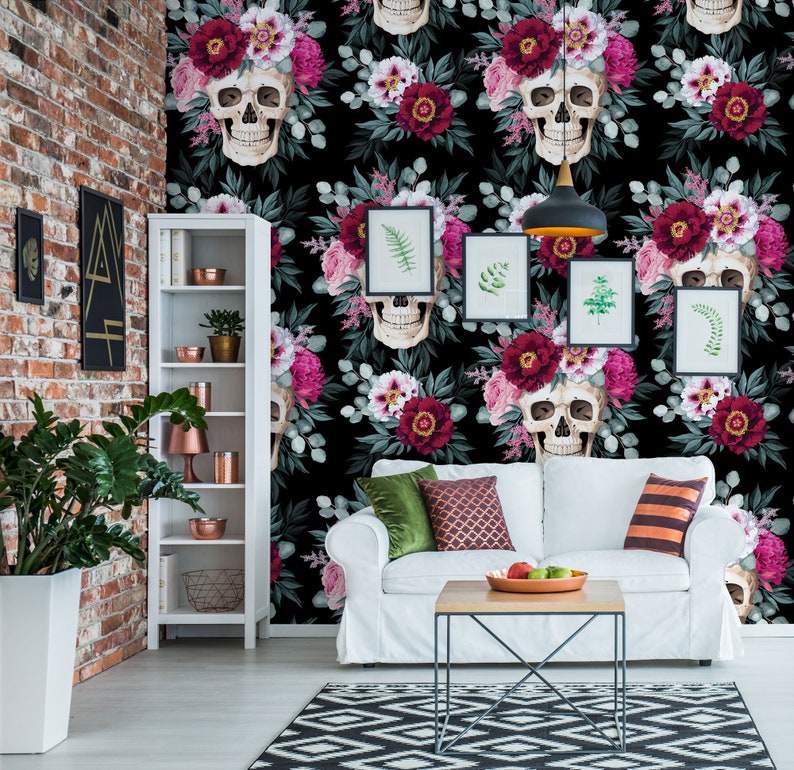 May include: A white couch with green and red pillows sits in front of a black and white floral wallpaper with skulls. The wallpaper features pink and red flowers, green leaves, and white skulls with floral crowns.
