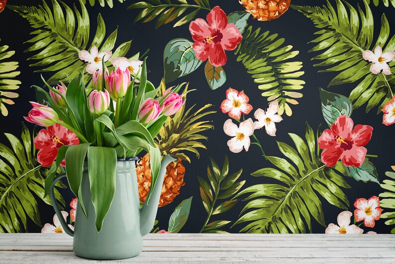 May include: A bouquet of pink tulips in a blue enamel pitcher against a backdrop of a dark blue floral wallpaper featuring tropical leaves, pineapples, and hibiscus flowers.