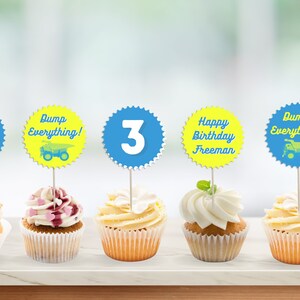 Puede incluir: Cinco cupcakes con glaseado azul y amarillo y adornos. Los adornos dicen "Happy Birthday Freeman", "Dump Everything!" con una imagen de un camión de volteo, "3", "Happy Birthday Freeman", y "Dump Everything!" con una imagen de un camión de volteo.