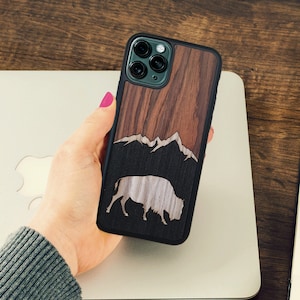 May include: A black and brown wood phone case with a mountain and bison design. The case has a cutout for the camera lens.