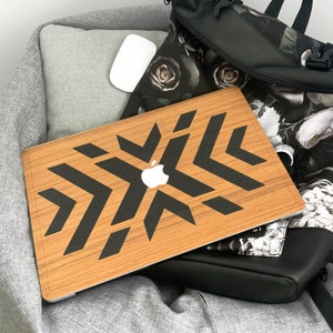 Puede incluir: Una funda de portátil de madera con un diseño geométrico negro. La funda tiene un patrón de copo de nieve con un logotipo de Apple negro en el centro.