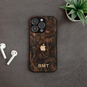 May include: A wooden phone case with a dark brown wood grain pattern and a gold Apple logo. The case has a black border and the initials "RMT" are engraved at the bottom. White wireless earbuds are on the left.