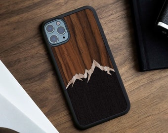 May include: A smartphone case featuring a black frame and a back panel with a wood grain inlay. The design displays a mountain range silhouette created by contrasting light and dark wood textures against a rich brown vertical grain background.