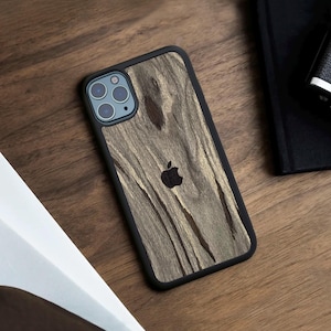 May include: A smartphone case with a black frame and a textured, light brown wood grain back panel. The case features a cutout for a triple-lens camera system and a centred black Apple logo. The design covers the back of the device completely.