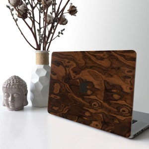 May include: A laptop computer with a wood grain design on the lid. The laptop is closed and sitting on a white surface. There is a small white vase with a cotton plant in it and a small gray Buddha statue on the surface.