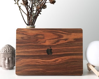 Puede incluir: Una funda para portátil de madera marrón con un logotipo de Apple negro. La funda está sobre una superficie blanca.