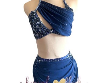May include: A two-piece dance costume in deep navy blue featuring a halter-style top with gathered fabric, a cutout detail, and dense crystal embellishments. The matching skirt has a draped overlay and crystal accents. Text reads burgundy & blush.