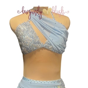May include: A light blue two-piece dance costume featuring a halter-style top with a draped fabric overlay and dense silver crystal embellishments. The matching skirt has a draped side panel with scattered crystals and a ruffled hemline.