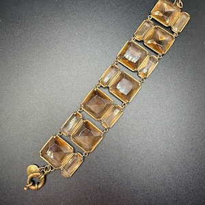 May include: A bracelet featuring alternating large square and smaller rectangular amber-colored faceted glass stones set in a brass-toned metal frame. The piece includes a circular toggle clasp with a small round metal tag attached to the end.