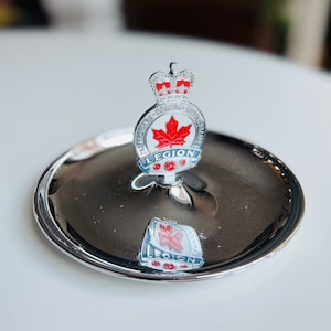 Puede incluir: Un porta anillos de metal plateado con un emblema de la Legión Real Canadiense. El emblema presenta una hoja de arce roja, una corona y las palabras "The Royal Canadian Legion".