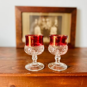 May include: Two clear glass wine goblets with a red and gold rim. The goblets have a cut glass design and are sitting on a wooden surface.