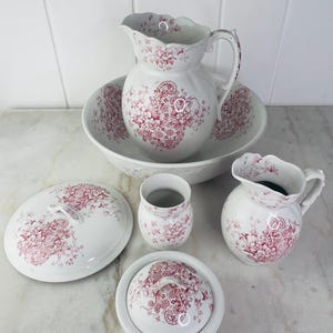 Może przedstawiać: Zestaw sześciu zabytkowych akcesoriów łazienkowych w kolorze białym z różowym wzorem kwiatowym. Zestaw zawiera dzbanek, miskę, słoik z pokrywką, talerz z pokrywką i dwa kubki.