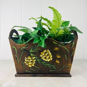 May include: A brown wooden planter with a wavy top edge and painted pine cone designs. The planter is filled with green leafy plants.