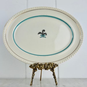 May include: A white oval serving platter with a gold rim and a teal blue border. The center of the platter features a black and teal design of a stylized leaf with a bow.