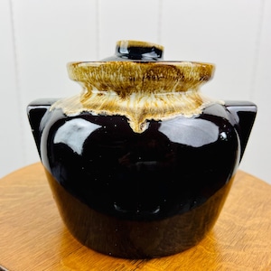 Könnte beinhalten: Ein brauner und cremefarbener, glasierter Keramiktopf mit Deckel. Der Topf hat zwei kleine Griffe an den Seiten.