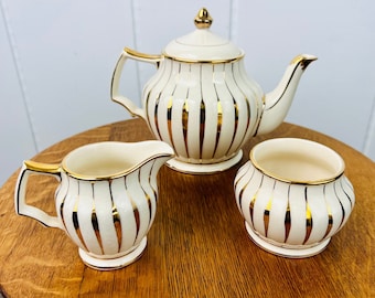 Puede incluir: Un juego de té de color crema que consta de una tetera, una jarra de leche y un azucarero. Cada pieza presenta una textura acanalada vertical con rayas doradas metálicas, asas con bordes dorados y bordes dorados. La tetera tiene una tapa a juego.