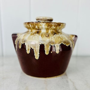 Könnte beinhalten: Ein kleiner brauner Keramiktopf mit einem weißen Überzug, der an den Seiten heruntertropft. Der Deckel hat einen braunen und weißen Überzug.