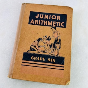 Peut inclure: Livre scolaire vintage "Junior Arithmetic" de sixième année. La couverture est beige avec du texte et des illustrations noirs. Le titre est dans un rectangle noir, avec une illustration de deux enfants nourrissant des poulets. Le livre est usé.