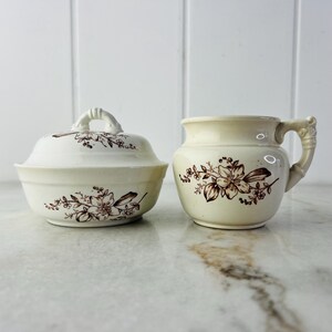 Op de afbeelding: Twee vintage keramische kommen met bruine bloemenmotieven. De kom links heeft een deksel. Beide kommen zijn wit met bruine bloemenmotieven.