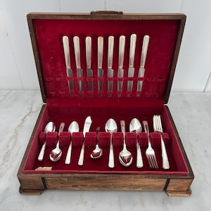 Puede incluir: Un estuche de cubiertos de madera con interior de terciopelo rojo, que contiene un juego completo de utensilios de plata. El juego incluye cuchillos, tenedores y cucharas, todos cuidadosamente dispuestos. El estuche está abierto, revelando el contenido.