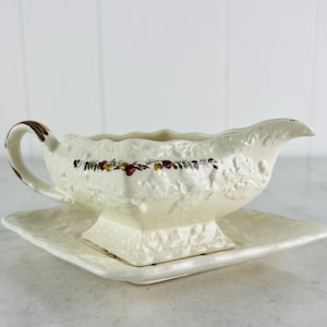 May include: A white ceramic gravy boat with a floral design and a matching saucer. The boat has a raised, textured surface and a gold rim.