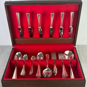 May include: A vintage silverware set presented in a wooden case with a red velvet lining. The set includes knives, spoons, forks, and serving utensils. The silverware has a shiny, silver finish. The case is open, revealing the neatly arranged pieces.