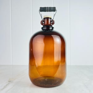 May include: A large, amber glass jug with a metal and wood handle. The jug has a round body and a narrow neck with a white cap. The handle is attached to the neck with metal wire. The jug is likely used for storing liquids.