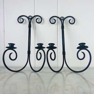 Peut inclure: Deux appliques murales en métal noir avec un motif en volutes. Chaque applique a une tige verticale avec un dessus décoratif et un bougeoir. Les appliques ont une base incurvée et sont conçues pour contenir des bougies. Les appliques sont en métal.