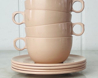 Puede incluir: Un juego de cuatro tazas de té y platillos de cerámica de color rosa pálido. Las tazas están apiladas una encima de la otra, y los platillos están apilados debajo.