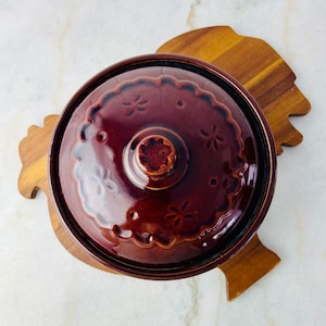 以下が含まれることがあります： 蓋と花柄のついた茶色の陶器製のキャセロール。鶏の形をした木製スタンドの上に置かれています。