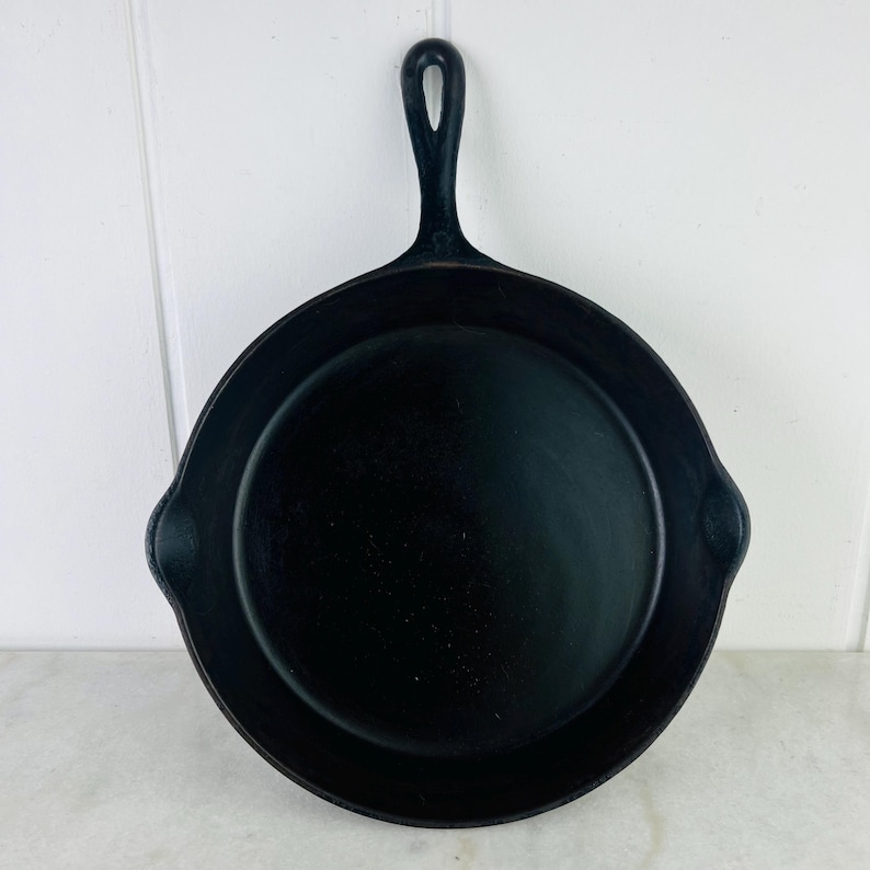 May include: A black cast iron skillet with a long handle and two small side handles. The skillet is round with a flat cooking surface and a raised edge. The handle has a hole for hanging. The skillet is made of heavy-duty metal.