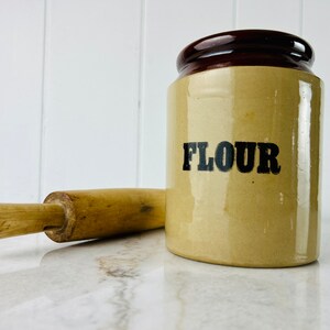 Peut inclure: Un pot à farine en céramique brun et beige avec un couvercle brun et le mot "FLOUR" imprimé en noir sur le devant. Un rouleau à pâtisserie en bois est au premier plan.
