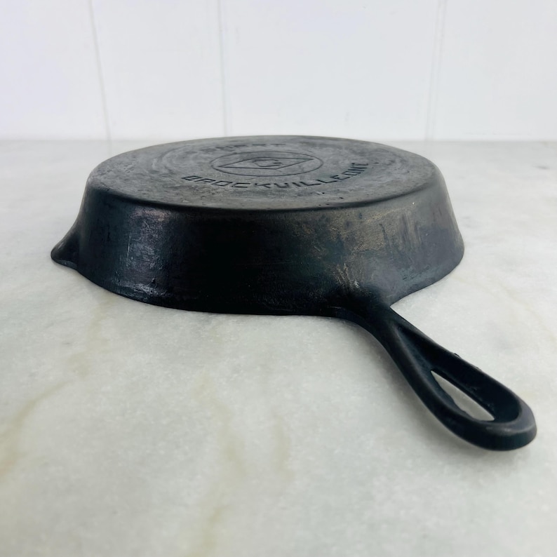 May include: Black cast iron skillet with a single handle. The skillet has a circular cooking surface and a slightly raised edge. The brand name "BROCKVILLE" is embossed on the bottom of the pan. The handle has a loop for hanging.