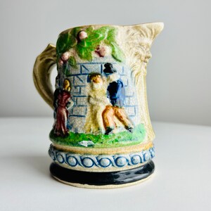 May include: A ceramic pitcher with a detailed scene of people in front of a brick wall. The pitcher has a handle and a base with a blue and white pattern.