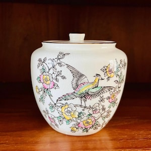 May include: White ceramic jar with a lid, featuring a colorful bird and floral design. The bird is depicted with outstretched wings and a long tail, surrounded by pink, yellow, and green flowers.