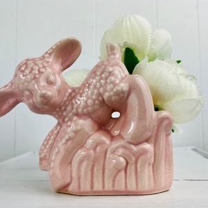 May include: A pink ceramic lamb planter with a textured surface. The lamb is sitting on a bed of green grass and has its head turned to the side. The planter is ideal for holding small plants or flowers.