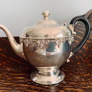 May include: A silver teapot with a black handle and a decorative grapevine design around the top. The teapot is sitting on a dark wooden surface.