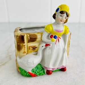 May include: A vintage ceramic planter featuring a woman in a yellow dress and bonnet, leaning against a wooden fence. A white goose stands on green grass. The planter has a rectangular shape and is painted with bright colors.
